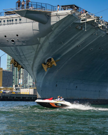 Small speedboat racing past the massive bow of a docked aircraft carrier at an urban harbor, high-rise skyline in the background.