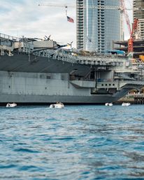 Massive gray aircraft carrier docked at an urban waterfront with skyscrapers and construction cranes behind it, an American flag flying from a crane and four small motorboats on the water in the foreground.