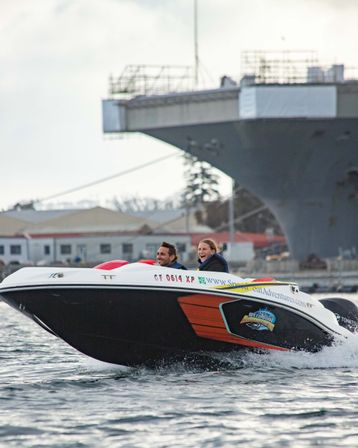 Two smiling passengers in a colorful speedboat skimming choppy harbor waters with a large gray naval ship and waterfront buildings in the background.