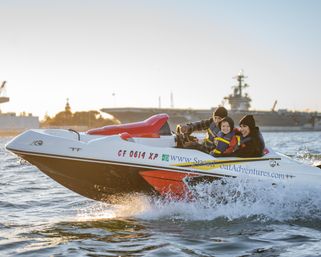 Family wearing life jackets on a red-and-white speedboat skimming a coastal harbor at golden hour, splashing water with a smiling child and a large naval ship and dock silhouette in the background.