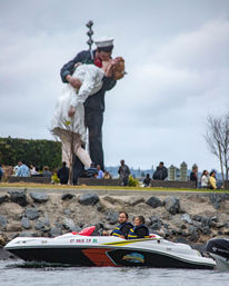 Couple in life jackets riding a small powerboat along a rocky San Diego waterfront, with a giant sailor-and-nurse kissing statue and people on the grassy promenade under a cloudy sky.