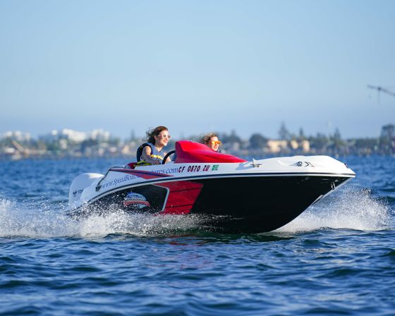 Two riders in life jackets and sunglasses enjoying a red-and-white speedboat skimming sparkling coastal waters near a harbor on a sunny day.