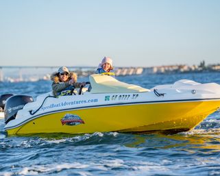 Two smiling people bundled in jackets and hats riding a bright yellow speedboat across a sunlit coastal bay, choppy blue water with a distant bridge and shoreline in the background.