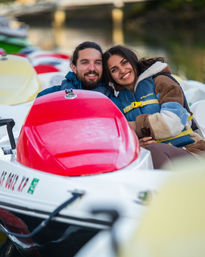 Smiling couple wearing life jackets cuddled in a red-topped pedal boat on a calm lake near a dock, enjoying a cozy waterfront outing.