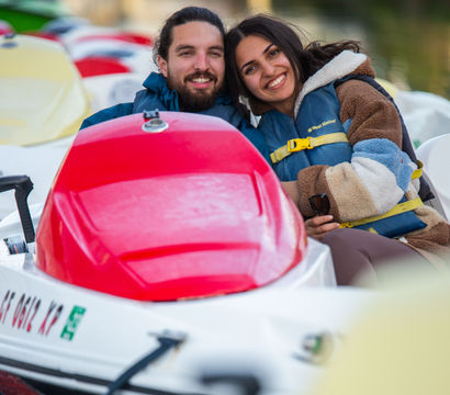 Smiling couple wearing life jackets cuddled in a red-topped pedal boat on a calm lake near a dock, enjoying a cozy waterfront outing.