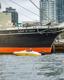 Yellow speedboat with a passenger in a life jacket passes the large black-and-orange hull of the tall ship 'Star of India' docked by waterfront high-rises.