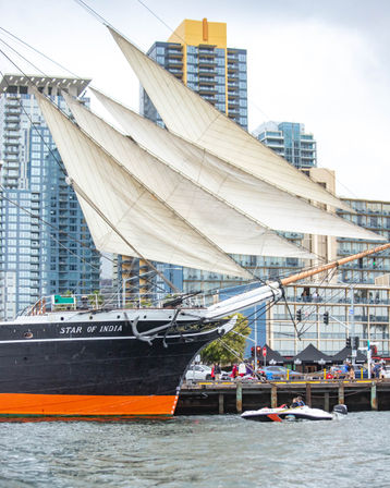 Historic tall ship 'Star of India' with cream sails unfurled, docked at an urban harbor against a backdrop of high-rise city skyline.