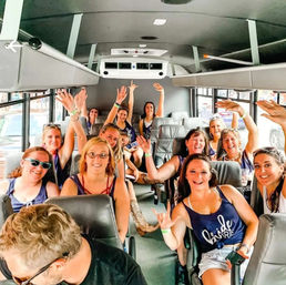 Cheerful bachelorette crew of women in matching tank tops smiling and waving inside a party bus interior