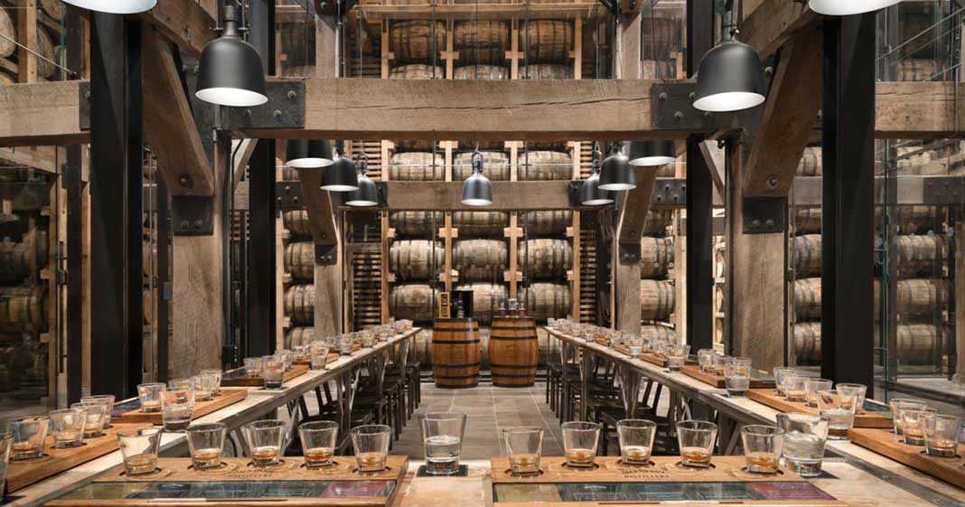 Rustic distillery tasting room inside a barrel warehouse — long communal wooden tables set with whiskey tasting flights, industrial pendant lights, and stacked oak barrels in a racked cellar.