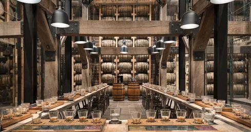 Rustic distillery tasting room inside a barrel warehouse — long communal wooden tables set with whiskey tasting flights, industrial pendant lights, and stacked oak barrels in a racked cellar.