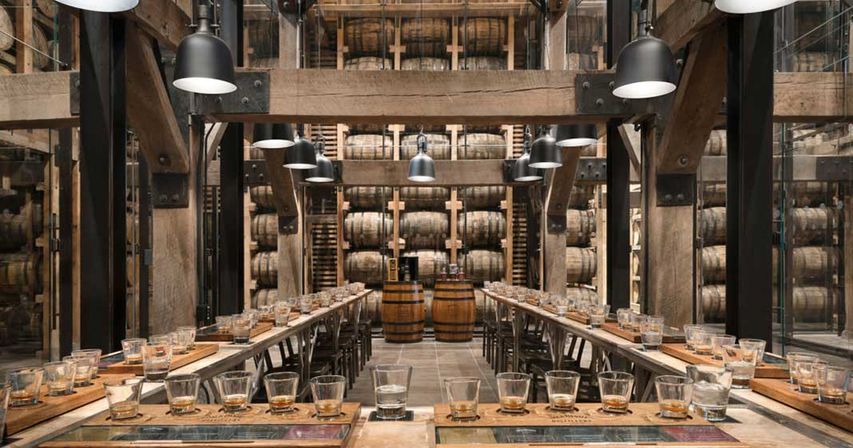 Rustic distillery tasting room inside a barrel warehouse — long communal wooden tables set with whiskey tasting flights, industrial pendant lights, and stacked oak barrels in a racked cellar.