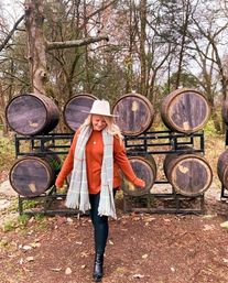 Woman in a white wide-brim hat, rust sweater, plaid scarf, black leggings and boots walking in front of stacked wooden oak barrels on metal racks in a rustic autumn woodland with fallen leaves.