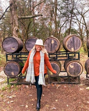 Woman in a white wide-brim hat, rust sweater, plaid scarf, black leggings and boots walking in front of stacked wooden oak barrels on metal racks in a rustic autumn woodland with fallen leaves.