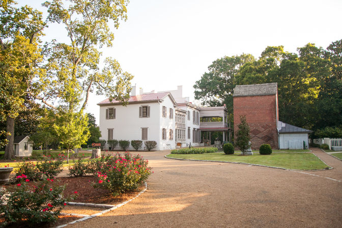 Sunlit white historic mansion with shuttered windows and a red-brick outbuilding, surrounded by rose beds, a gravel circular driveway and mature shade trees in a landscaped estate