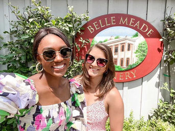 Two smiling women in sunglasses pose outdoors in front of a red winery sign and leafy vines on a sunny day