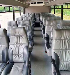 Bright shuttle bus interior with rows of plush gray leather seats, central aisle and large windows framing green roadside scenery.