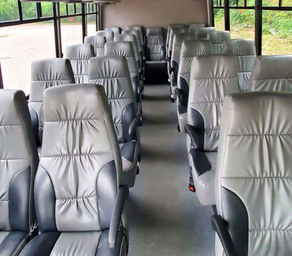 Coach bus interior with empty rows of gray leather reclining seats and armrests, a central aisle and large windows showing green foliage outside.