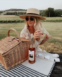 Woman in a straw hat and round sunglasses sipping rosé at a picnic in a vineyard, wicker picnic basket and wine bottle on a striped blanket with rolling green hills in the background.