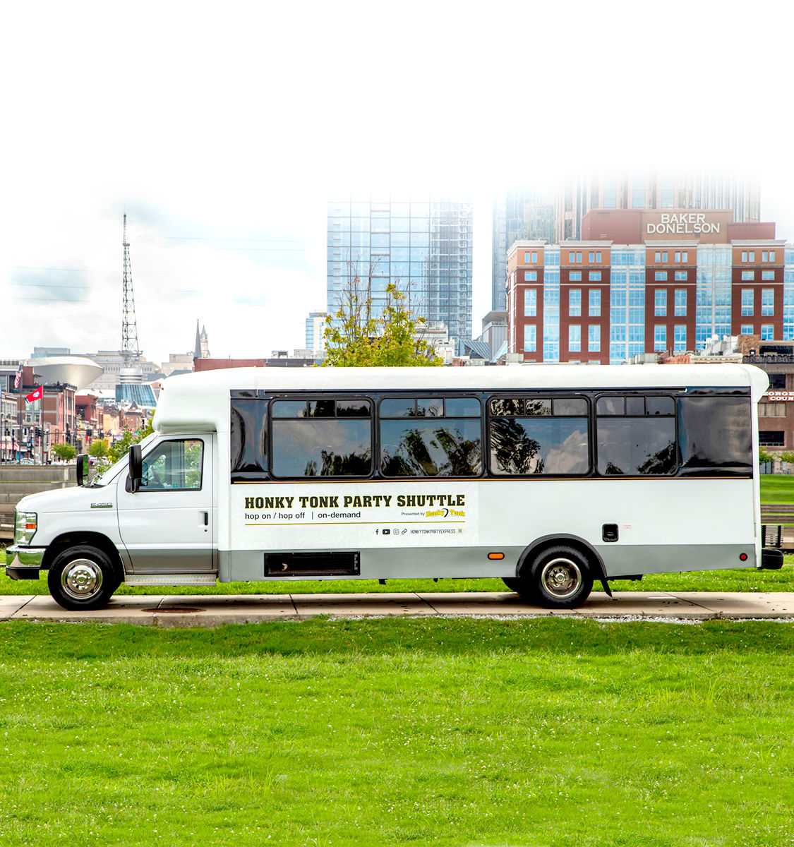 White party shuttle bus parked beside a green lawn with a downtown city skyline of glass and brick high-rise buildings in the background.