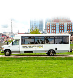 White party shuttle bus parked beside a green lawn with a downtown city skyline of glass and brick high-rise buildings in the background.