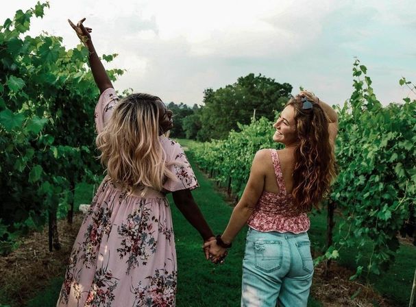 Two friends holding hands walking down a lush vineyard row, one in a floral dress and the other in a pink top and jeans, smiling in a countryside wine-country setting.