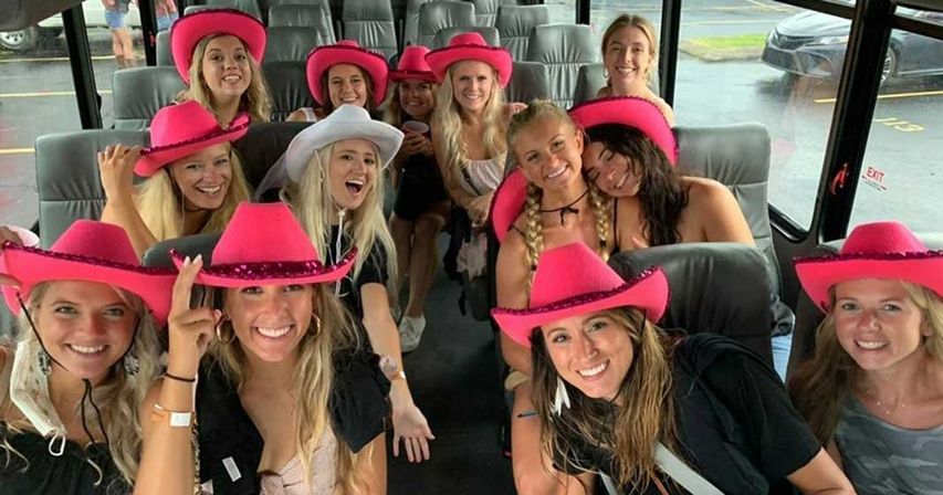 Group of smiling women in bright pink (and one white) cowgirl hats posing inside a party bus with leather seats, parked in a rainy parking lot — festive bachelorette or girls’ outing vibe.