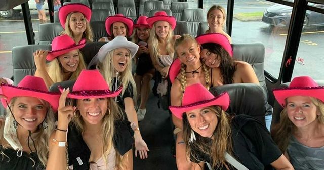 Group of smiling women in bright pink (and one white) cowgirl hats posing inside a party bus with leather seats, parked in a rainy parking lot — festive bachelorette or girls’ outing vibe.