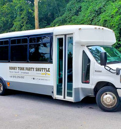 White party shuttle bus with sliding glass doors and tinted windows, labeled for honky-tonk style party service, parked on a tree-lined street.