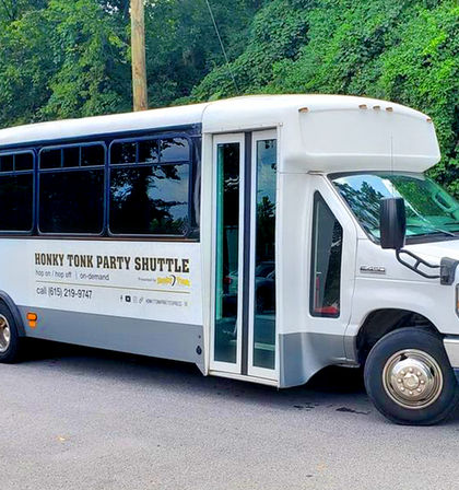 White party shuttle bus with sliding glass doors and tinted windows, labeled for honky-tonk style party service, parked on a tree-lined street.