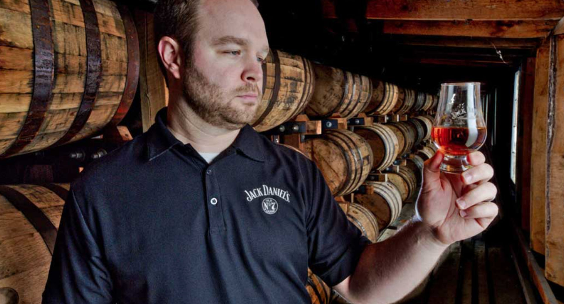 Man inspecting a glass of amber whiskey in a distillery barrel room lined with stacked oak casks
