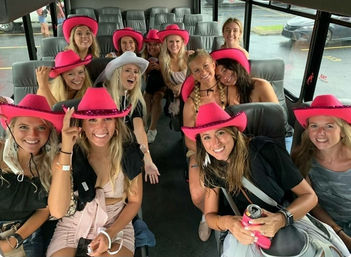Cheerful group of women wearing bright pink cowboy hats and casual summer outfits, laughing and posing together inside a party bus with gray seats and rainy windows visible outside.
