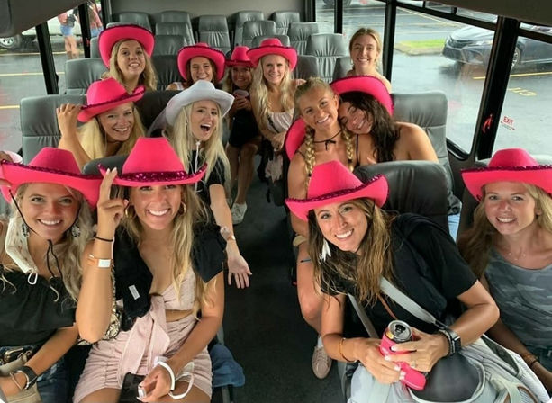 Cheerful group of women wearing bright pink cowboy hats and casual summer outfits, laughing and posing together inside a party bus with gray seats and rainy windows visible outside.