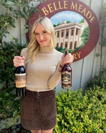 Smiling woman holding a bottle of red wine and a bottle of bourbon in front of a painted distillery sign and garden foliage