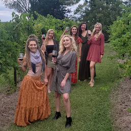 Six women in colorful dresses smiling and raising wine glasses while strolling a lush vineyard row
