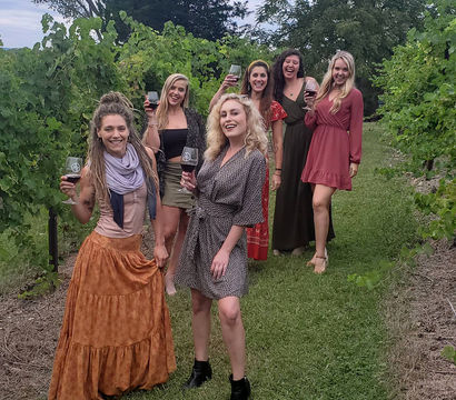 Six women in colorful dresses smiling and raising wine glasses while strolling a lush vineyard row