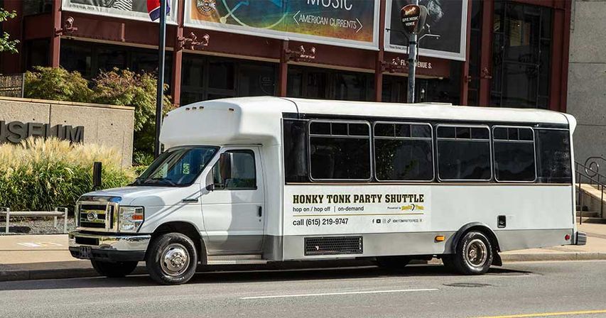 White party shuttle bus with tinted windows parked on a downtown street in front of a museum, advertising hop-on hop-off nightlife tours