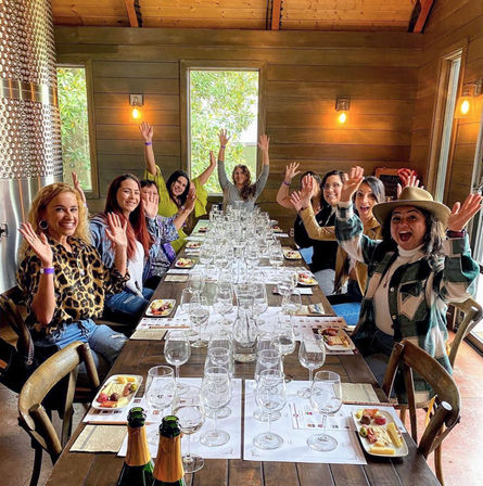 Group of friends waving around a long wooden table set for wine tasting in a rustic winery tasting room with rows of wine glasses, cheese plates, and champagne bottles.