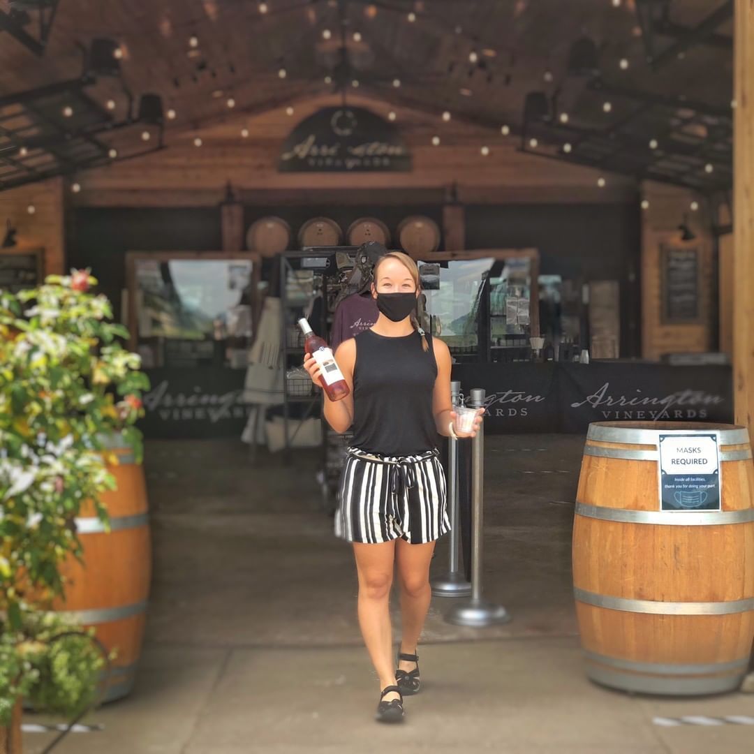 Masked server carrying a bottle of rosé and a tasting cup walking out of a winery tasting room between wooden barrels and string lights.