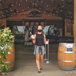 Masked server carrying a bottle of rosé and a tasting cup walking out of a winery tasting room between wooden barrels and string lights.