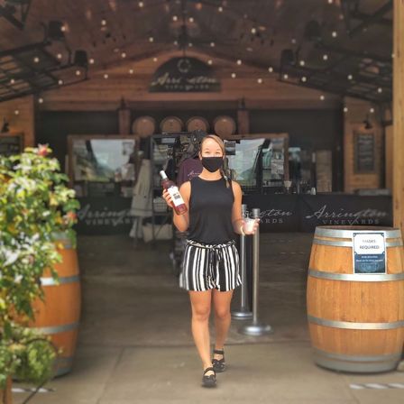 Masked server carrying a bottle of rosé and a tasting cup walking out of a winery tasting room between wooden barrels and string lights.