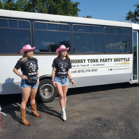 Two women in pink cowboy hats, matching 'bridesmaid' tees, denim shorts and cowboy boots posing beside a white country-style party shuttle bus on a sunny day.
