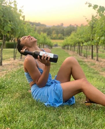Joyful person in a blue dress reclining on grass and holding a bottle of red wine among vineyard rows at golden sunset