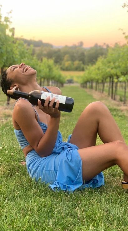 Joyful person in a blue dress reclining on grass and holding a bottle of red wine among vineyard rows at golden sunset