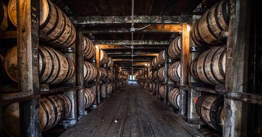 Dim wooden distillery warehouse interior with a long center aisle lined on both sides by stacked oak barrels aging spirits, warm light glowing at the far end.
