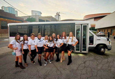 Group of women in matching white tees and black skirts posing in front of a white party bus on a downtown street at dusk, city buildings visible — girls night out / bachelorette party vibe.