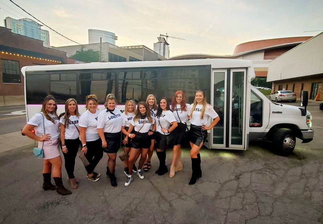 Group of women in matching white tees and black skirts posing in front of a white party bus on a downtown street at dusk, city buildings visible — girls night out / bachelorette party vibe.