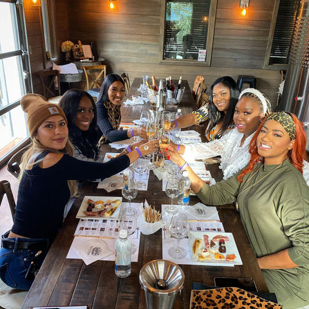 Six women clinking wine glasses over charcuterie plates at a rustic winery tasting room table with wooden walls and daylight