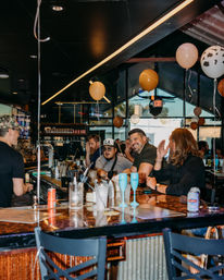 Lively urban bar nightlife: friends laughing at a busy counter with balloons, cocktails, draft taps and a bartender.
