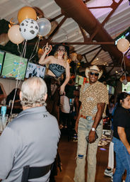 Lively cowboy-themed sports bar scene with balloons, woman in a bandana top standing on a table, smiling man in a cowboy hat and leopard shirt, and patrons watching TVs.