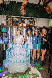 Country-themed indoor bar party with four women in cowboy hats and boots; one wears a tiered tulle dress displaying dozens of light-blue champagne flutes, with beer kegs, a mounted zebra head and a 'YEE HAW' sign in the background.